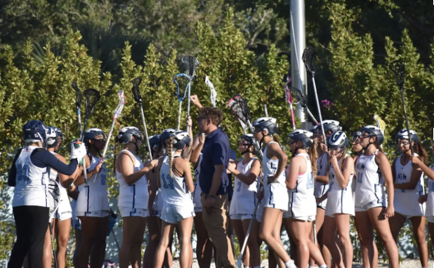 Members of the Girls Lacrosse team huddling up before a game, alongside their coach.